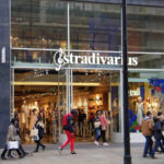 Two young women exit an upscale clothing store as nine pedestrians pass by. Large windows with the word Stradivarius painted on them expose the posh fashions inside.