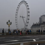 Under an overcast gray sky, people stand in line for the London Eye ferris wheel, which stands next to a light colored two-story building with a dark roof.
