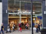 Two young women exit an upscale clothing store as nine pedestrians pass by. Large windows with the word Stradivarius painted on them expose the posh fashions inside.